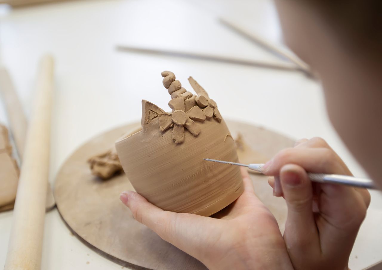 Dłonie dziecka rzeźbiące narzędziem gliniany kubek w pracowni ceramicznej